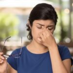 A young woman with dark hair and a blue shirt holds glasses and pinches the bridge of her nose, appearing stressed or having a headache.